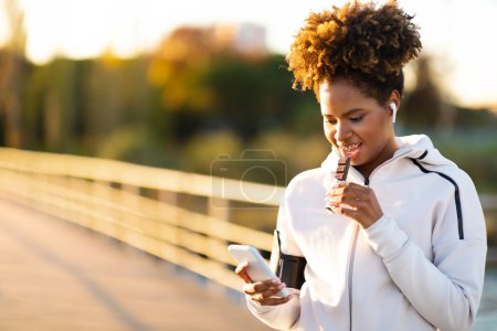 Photo for Sporty Young Black Woman Having Rest After Outdoor Training, Eating Protein Bar And Using Smartphone, African American Female Athlete Browsing Fitness Tracker App And Enjoying Snack, Copy Space - Royalty Free Image