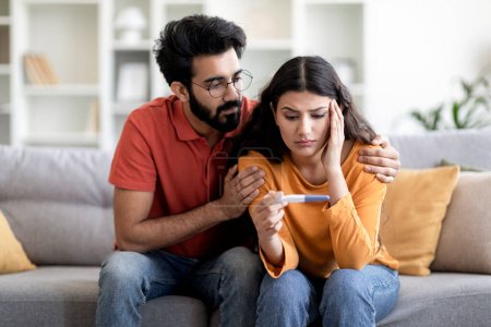 Photo for Infertility Problems. Portrait Of Upset Young Indian Couple Looking At Negative Pregnancy Test While Sitting Together On Couch At Home, Caring Eastern Husband Comforting Depressed Wife, Free Space - Royalty Free Image