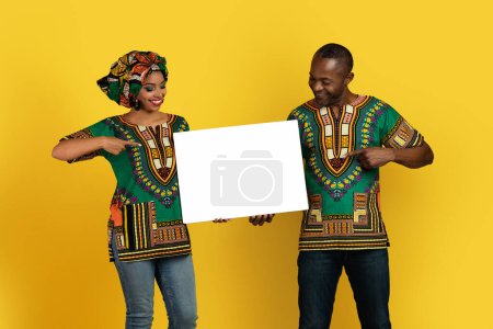Foto de Emocional alegre feliz hermosa pareja negra en trajes africanos tradicionales coloridos apuntando a la pizarra blanca en blanco con maqueta para publicidad o texto, fondo amarillo del estudio - Imagen libre de derechos