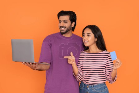 Photo for Cheerful beautiful loving young indian couple in casual holding modern pc laptop and blue plastic bank card, woman pointing at computer screen, lovers booking tickets online, orange background - Royalty Free Image