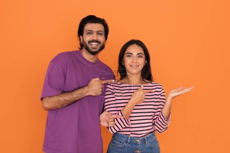 Téléchargez les photos : Amour joyeux beau jeune couple indien dans casual hindou homme et femme pointant vers l'espace libre au-dessus de la main féminine, souriant à la caméra, isolé sur fond de studio orange - en image libre de droit