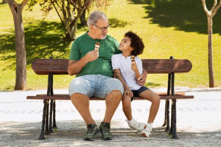 Téléchargez les photos : Souriant vieux grand-père européen et métis petit garçon manger de la crème glacée dans le parc sur le banc, profiter du temps libre ensemble, en plein air. Bonbons, week-end en famille en ville en été, amour et relation - en image libre de droit
