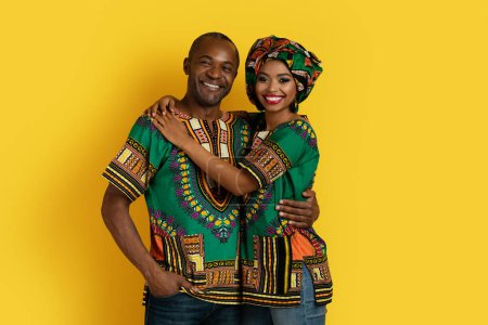 Foto de Alegre feliz amante marido negro y esposa en hermosos trajes africanos tradicionales posando juntos en el fondo del estudio amarillo, abrazos, sonriendo a la cámara. Amor, matrimonio, relaciones - Imagen libre de derechos