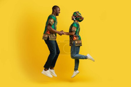 Photo for We getting married. Emotional happy african couple wearing traditional bright costumes holding hands and jumping up over yellow studio background, celebrating good news, copy space - Royalty Free Image