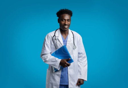 Téléchargez les photos : Joyeux beau jeune homme noir médecin posant sur fond bleu studio, vêtu d'un manteau médical blanc, stéthoscope suspendu, dossier de maintien avec anamnèse du patient, espace de copie - en image libre de droit
