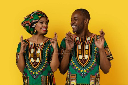 Photo for Loving attractive adult black couple in bright national african clothing posing on yellow studio background, holding fingers crossed and looking at each other, believe in superstitions, good luck - Royalty Free Image