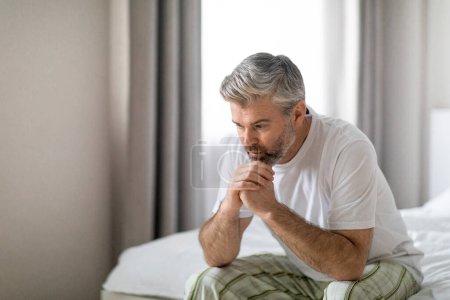 Photo for Depressed middle aged grey-haired handsome man wearing pajamas sitting on bed alone at home, leaning on his hands, suffering from loneliness or anxiety, copy space. Permacrisis, midlife crisis - Royalty Free Image