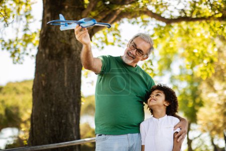 Photo for Cheerful elderly european grandfather and mixed race little boy play toy airplane in park, enjoy free time together, outdoor. Dreaming, family hobby at weekend, entertainment - Royalty Free Image