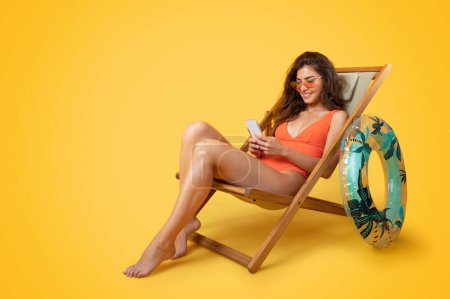 Foto de Sonriente milenaria mujer asiática en traje de baño, gafas de sol con anillo inflable sentarse en la silla de cubierta, disfrutar del tiempo libre, charlando en el teléfono aislado sobre fondo naranja. Verano con gadget, descanso y relax - Imagen libre de derechos