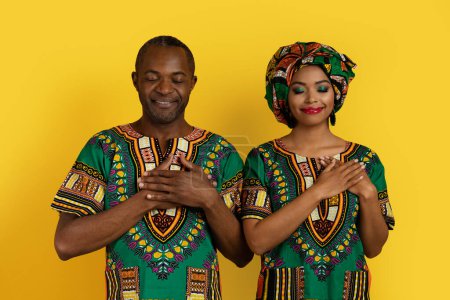 Photo for Peaceful smiling african american lovers middle aged man and young woman in traditional costumes holding hands on chest over heart, expressing love and affection, yellow studio background - Royalty Free Image