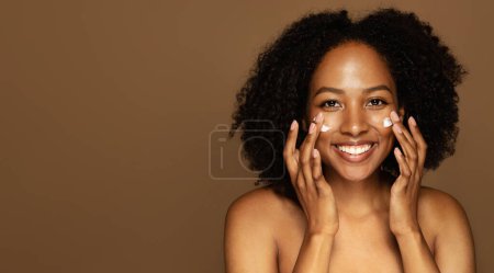 Téléchargez les photos : Soins de la peau. Gros plan studio de joyeux attrayant topless africaine américaine millénaire femme massant le visage avec les doigts et souriant à la caméra, appliquer de la crème, isolé sur brun, espace de copie - en image libre de droit