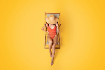 Téléchargez les photos : Femme asiatique millénaire en maillot de bain et chapeau dormir sur chaise longue, profiter du temps libre, isolé sur fond de studio orange, vue de dessus, méconnaissable. Annonce et offre en été, repos et détente - en image libre de droit