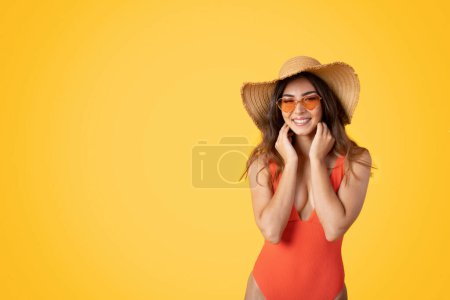 Foto de Sonriente milenaria mujer asiática en traje de baño, sombrero y gafas de sol disfruta de vacaciones en el complejo, viaje a la playa, aislado en el fondo del estudio naranja. Vacaciones en verano, descanso y relax, turismo - Imagen libre de derechos