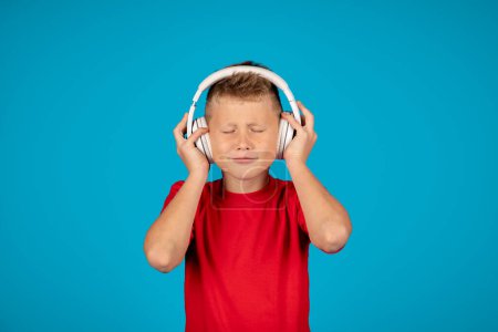 Foto de Retrato de niño divertido escuchando música en auriculares inalámbricos, niño preadolescente alegre de pie con los ojos cerrados, disfrutando de canciones favoritas mientras posan sobre fondo azul, espacio de copia - Imagen libre de derechos