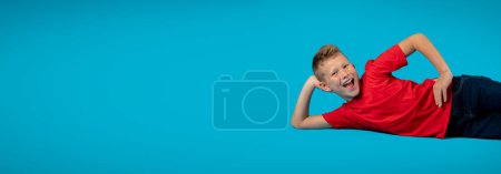 Photo for Cheerful Male Child Relaxing On Floor While Posing Isolated On Blue Background, Creative Banner With Positive Preteen Boy Smiling At Camera, Joyful Kid Having Fun In Studio, Panorama With Copy Space - Royalty Free Image