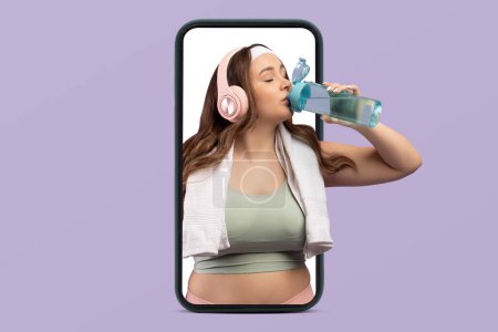 Photo for Thirsty young caucasian woman plus size in sportswear with headphones drink bottle of water on phone screen isolated on violet studio background. Music for workout, body care app, weight loss - Royalty Free Image