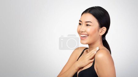 Photo for Bodycare Concept. Joyful Asian Woman Looking Aside At Free Space Touching Shoulders And Radiant Skin Posing Over Gray Studio Background. Skincare And Beauty Products Advertisement. Panorama - Royalty Free Image