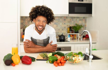 Téléchargez les photos : Cuisine et recettes. Gai homme afro-américain posant à la table de la cuisine avec des légumes sur le dessus à la maison, souriant à la caméra. Portrait de chef portant un tablier. Préparation saine des repas et des dîners - en image libre de droit