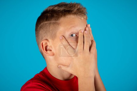 Photo for Shy preteen boy hiding face and peeking out through fingers, curious tween male child looking at camera with interest, caucasian kid posing isolated over blue studio background, copy space - Royalty Free Image