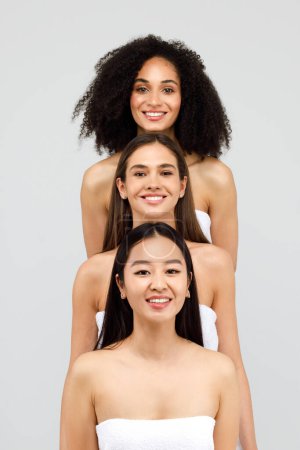Foto de Grupo de tres mujeres hermosas diversas sonriendo a la cámara, de pie y posando sobre fondo gris estudio. Retrato de belleza vertical de damas jóvenes - Imagen libre de derechos