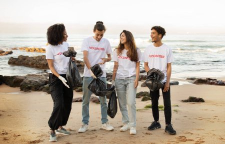 Téléchargez les photos : Des jeunes gens joyeux et divers bénévoles dans des t-shirts avec des sacs à ordures nettoient les ordures sur la plage de la mer, s'amusent en plein air, sur toute la longueur. Eco conservation, environnement, protection de la planète - en image libre de droit