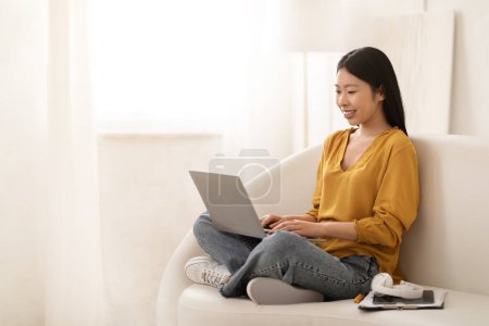 Photo for Positive smiling pretty young asian woman wearing casual outfit sitting on couch in living room, working from home, using laptop, chinese lady freelancer typing on computer keyboard, copy space - Royalty Free Image