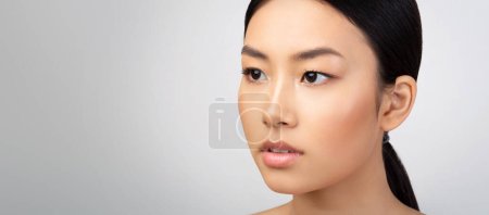 Téléchargez les photos : Portrait de beauté de dame asiatique attrayante regardant à côté de l'espace libre sur fond de studio gris clair. Gros plan de jeunes femmes visage avec peau lisse parfaite et maquillage. Panorama - en image libre de droit