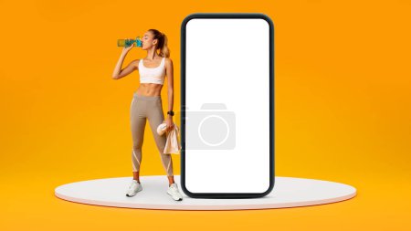 Photo for Tired young european lady in sportswear with towel resting, drink bottle of water near big smartphone with empty screen isolated on orange studio background, panorama. Aqua balance, app for sports - Royalty Free Image