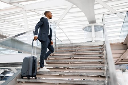 Foto de Joven hombre de negocios negro con maleta que sube escaleras en la terminal del aeropuerto, Handsoem afroamericano Millennial masculino que usa traje que va a la puerta de salida del vuelo, teniendo viaje de negocios, espacio de copia - Imagen libre de derechos