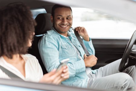 Téléchargez les photos : Circuit sûr en voiture. heureux indien conducteur homme fixation ceinture de sécurité sourire à passager dame comme elle utilise téléphone portable assis dans l'automobile à l'intérieur. Propriété et transport de véhicules. Focus sélectif - en image libre de droit