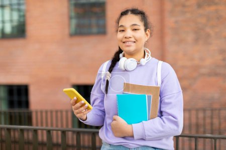 Téléchargez les photos : Joyeuse jeune femme métissée avec sac à dos, livres et smartphone debout sur le campus de l'université, marchant à l'extérieur après les cours. App pour l'étude, la connaissance, l'éducation et la leçon - en image libre de droit