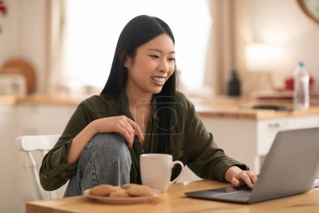 Photo for Happy beautiful young asian woman in casual relaxing in kitchen, sitting at desk in front of laptop, drinking coffee, eating cookies while watching movie or video content on Internet, copy space - Royalty Free Image