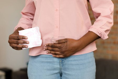 Téléchargez les photos : Lutte contre les douleurs menstruelles. Dame noire tenant une serviette hygiénique à la main, souffrant de règles douloureuses ou de crampes abdominales, serrant l'estomac avec une douleur menstruelle sévère - en image libre de droit