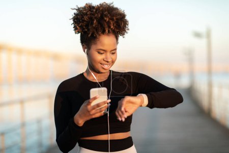 Photo for Fitness Tracker. Sporty black woman looking at smartwatch after training outdoors, smiling african american female holding smartphone, using modern gadgets for checking sport activity, copy space - Royalty Free Image