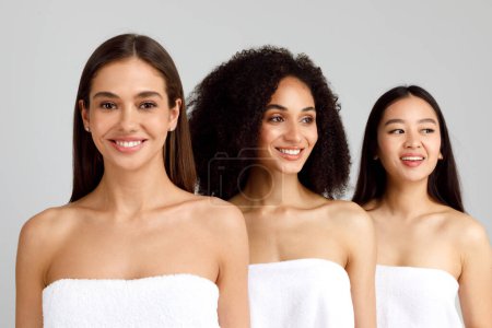 Foto de Tres jóvenes mujeres multirraciales felices posando en toallas de baño blancas, de pie en la fila y sonriendo sobre fondo gris, filmado en el estudio. Emocionado señora caucásica mirando a la cámara - Imagen libre de derechos