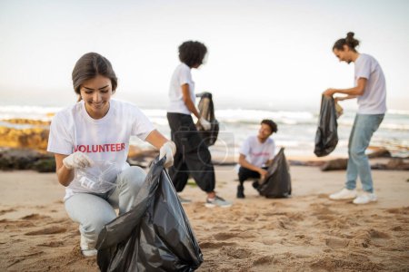 Téléchargez les photos : Des jeunes volontaires internationaux positifs dans des gants et une dame européenne avec des sacs poubelles nettoient les ordures sur la plage, en plein air. Conservation de l'environnement et de l'environnement protection de la planète, pollution des océans - en image libre de droit