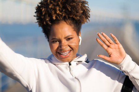 Téléchargez les photos : Gros plan de belle jeune femme noire en vêtements de sport prenant Selfie à l'extérieur, souriante afro-américaine avec des appareils dentaires agitant à la caméra tout en faisant des photos sur la formation à l'extérieur - en image libre de droit