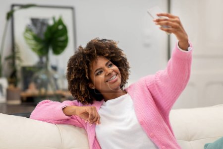 Téléchargez les photos : Heureuse jolie femme noire millénaire aux cheveux touffus assise sur le canapé seule, prenant des photos selfie, utilisant le téléphone à la maison, souriant à l'écran du smartphone, intérieur confortable du salon, espace de copie - en image libre de droit
