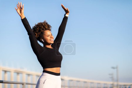 Foto de Estilo de vida activo. Mujer negra feliz en ropa deportiva disfrutando del ejercicio al aire libre, sonriendo a una joven afroamericana con auriculares levantando las manos y mirando la luz del sol, estirándose fuera, Copiar el espacio - Imagen libre de derechos