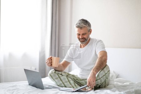 Photo for Happy handsome middle aged businessman wearing pajamas sitting on bed at home or hotel room, drinking coffee in the morning, using laptop, checking his planner notebook, copy space - Royalty Free Image