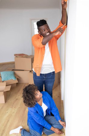 Foto de Jóvenes cónyuges negros midiendo paredes en su nuevo apartamento después de mudarse, feliz pareja afroamericana tomando medidas en la habitación, preparándose para la reparación y renovación, tiro vertical - Imagen libre de derechos