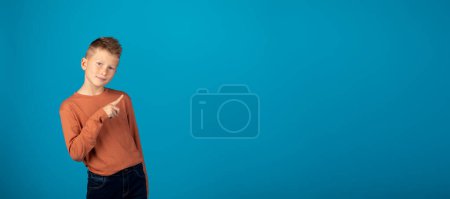 Photo for Check This. Smiling Caucasian Boy Pointing Aside At Copy Space With Finger, Cute Preteen Male Child Showing Free Place For Advertisement While Standing Over Blue Background, Panorama - Royalty Free Image