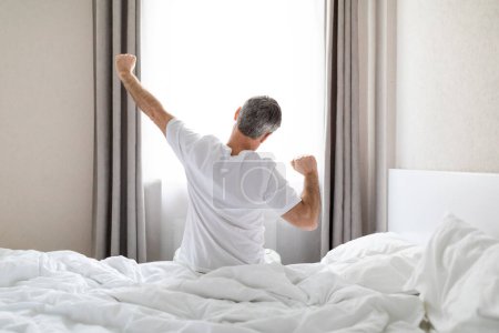 Back view of unrecognizable grey-haired man in pajamas sitting on bed and stretching body after waking up in the morning, looking at window, copy space. Comfortable healthy sleep concept