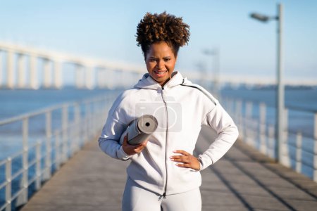 Téléchargez les photos : S'adapter à l'entraînement d'athlète féminine noire en plein air, tenant un tapis de fitness et regardant la caméra, attrayant jeune femme afro-américaine en vêtements de sport debout sur une jetée en bois près de la mer, s'exerçant à l'extérieur - en image libre de droit