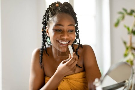 Beautiful young black woman using moisturizing lipbalm, looking at small mirror while sitting at vanity table and applying everyday nude makeup at home, copy space