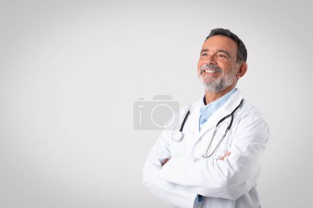 Photo for Smiling european senior man doctor in white coat with a stethoscope with crossed arms on chest, enjoy work, look at copy space on gray background. Medical care, diagnostics, exam, ad and offer - Royalty Free Image