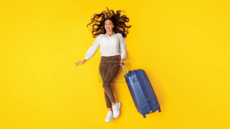 Téléchargez les photos : Offre de voyage. Joyeux jeune femme posant avec une valise bleue agence de voyage publicitaire couché sur fond jaune, vue de dessus. Plan complet de la femme touristique avec des bagages. Concept de vacances. Panorama - en image libre de droit