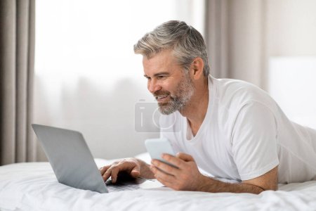Foto de Feliz guapo de pelo gris barbudo hombre de mediana edad nómada digital con pijamas acostado en la cama con teléfono celular y portátil, espacio de copia vista lateral. Concepto de tecnologías modernas - Imagen libre de derechos