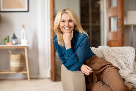 Photo for Positive Blonde Lady Smiling To Camera Enjoying Mature Life, Posing On Cozy Couch Resting Head On Fist And Expressing Happiness At Home. Elegant Senior Woman Relaxing In Living Room On Weekend - Royalty Free Image