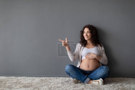 Foto de Anuncio de embarazo. Sonriendo embarazada mujer señalando a un lado en el espacio de copia en la pared gris mientras se relaja en el piso en casa, hermosa mujer embarazada abrazando el vientre y mostrando antecedentes para la publicidad - Imagen libre de derechos